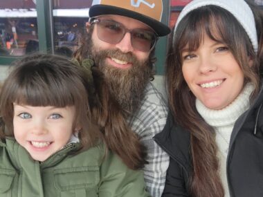 A married couple is pictured with one of their daughters. All are bundled up in coats and warm clothes. The man and woman are seated next to each other, and the girl, maybe about 5 or 6, looks to be sitting on her dad's lap.