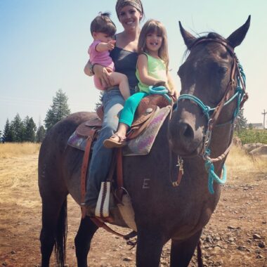 A woman, perhaps in her 20s, sits atop a brown horse in a field. She's wearing jeans and a black top, and her hair is tied back under some sort of headband or scarf. One of her daughters, maybe about 3 years old, sits in front of her on the saddle, while the woman holds her younger daughter, maybe about a year old, in her right arm. There are some tall green trees in the distance, but the field is mostly dirt, rocks, and some yellowed grass.