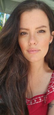 A close-up photo, which appears to be a selfie, shows a woman with long brown hair wearing a red shirt with white stitching. She looking at the camera intently and looks to be sitting on some sort of covered patio outside. Palm trees and a bit of blue sky are just visible in the background.