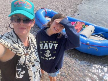 A woman and child stand along a shoreline in front of an inflatable raft.
