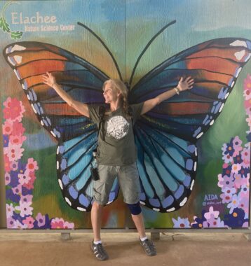 A woman stands with her arms outstretched in front of a large butterfly that's been painted on the wall.
